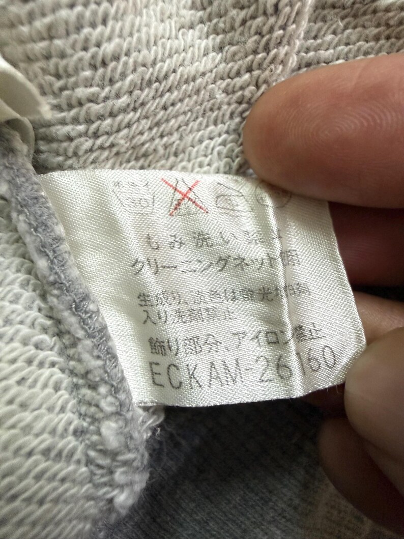 May include: Close-up of a clothing tag with Japanese text and the number ECKAM-26160. The tag is white with black text and a red symbol. The fabric is a light gray, textured material.