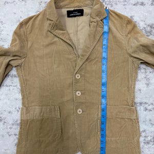 Puede incluir: Una blazer de pana color beige con cierre de tres botones, dos bolsillos delanteros y solapa con muescas. El blazer tiene una etiqueta que dice "tricot COMME des GAR&Ccedil;ONS". El blazer mide aproximadamente 66 cm de largo.