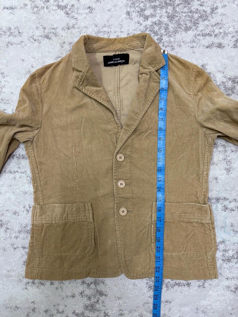 Puede incluir: Una blazer de pana color beige con cierre de tres botones, dos bolsillos delanteros y solapa con muescas. El blazer tiene una etiqueta que dice "tricot COMME des GAR&Ccedil;ONS". El blazer mide aproximadamente 66 cm de largo.