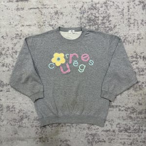 May include: A gray crew neck sweatshirt with the word "courage" in colorful, embroidered letters and a yellow flower graphic. The sweatshirt has long sleeves and a ribbed hem and cuffs. The fabric appears to be a soft, comfortable material.