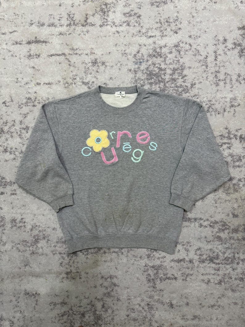 May include: A gray crew neck sweatshirt with the word "courage" in colorful, embroidered letters and a yellow flower graphic. The sweatshirt has long sleeves and a ribbed hem and cuffs. The fabric appears to be a soft, comfortable material.
