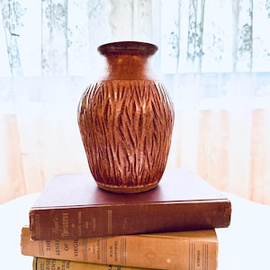 May include: A brown ceramic vase with a textured, vertical pattern, sitting atop a stack of three vintage books. The books are various shades of brown and tan. The background features a sheer curtain.