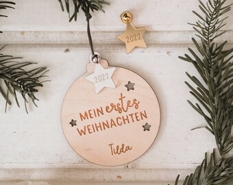 May include: A wooden Christmas ornament with the text "Mein erstes Weihnachten" and the name "Tilda". The ornament is round with star cutouts and a star-shaped accent with the year "2022". A small gold star-shaped bell is also present.