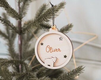 May include: A wooden Christmas ornament in the shape of a circle with the name "Oliver" engraved in a cursive font. The ornament features a bear and star cutouts. It hangs from a black string and is displayed on a Christmas tree.