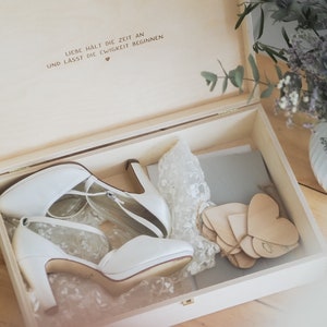 May include: A wooden box with a lid open, revealing a pair of white bridal shoes, a lace fabric, and a stack of wooden heart-shaped tags. The box has a handwritten inscription on the inside of the lid that reads "Liebe h&auml;lt die Zeit an und lasst die Ewigkeit beginnen" with a small heart symbol.