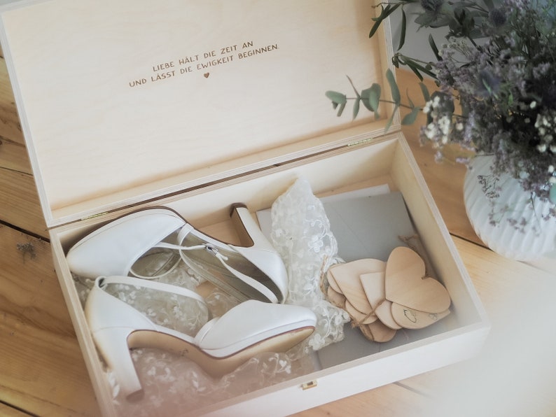 May include: A wooden box with a lid open, revealing a pair of white bridal shoes, a lace fabric, and a stack of wooden heart-shaped tags. The box has a handwritten inscription on the inside of the lid that reads "Liebe h&auml;lt die Zeit an und lasst die Ewigkeit beginnen" with a small heart symbol.