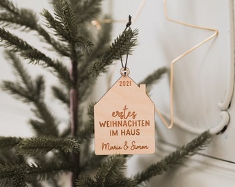 May include: A wooden house-shaped Christmas ornament with the year 2021 engraved at the top. The ornament has the words "erstes WEIHNACHTEN IM HAUS Marie & Simon" engraved on it. The ornament is hanging on a Christmas tree.