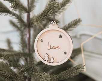 May include: A wooden Christmas ornament in the shape of a ball, hanging from a tree. The ornament has the name "Liam" engraved on it, along with star and tree cutouts. The ornament is made of light-colored wood.