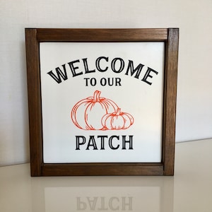 Peut inclure: Une pancarte en bois encadrée avec le texte "Welcome to our Patch" en lettres noires. Deux citrouilles orange sont représentées sous le texte.