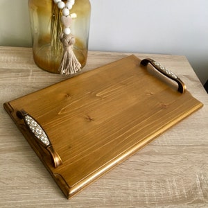 Peut inclure: Un plateau de service rectangulaire en bois avec deux poignées en métal ornées. Les poignées ont un motif blanc et or. Le plateau est teinté d'une couleur brun foncé.
