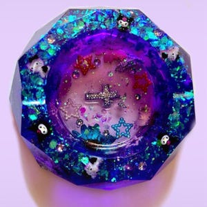 Può includere: Un posacenere ottagonale in resina nei toni del blu e del viola intenso, con glitter iridescenti incorporati, piccoli ciondoli a forma di stella, minuscoli volti di personaggi e una croce argentata al centro. La superficie è sfaccettata.