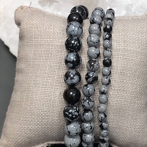 Peut inclure: Quatre bracelets en perles noires et grises empilés sur un coussin en tissu beige. Les bracelets sont faits de perles rondes et mouchetées.