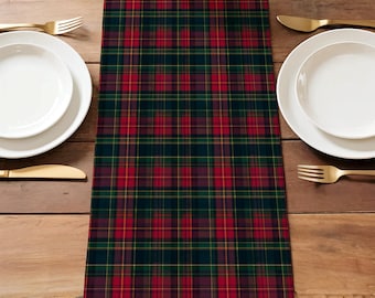 May include: A rectangular table runner with a classic plaid pattern in shades of red, green, and gold. The runner is placed on a wooden table, with white plates and gold cutlery on either side. A woven basket sits at the top.