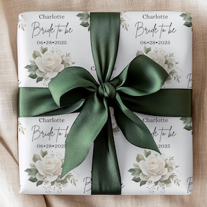 Peut inclure: Papier cadeau blanc avec un ruban de satin vert. Le papier a un motif r&eacute;p&eacute;t&eacute; de roses blanches et de feuilles vertes. Le texte "Charlotte Bride to be 06.28.2025" est imprim&eacute; sur le papier.