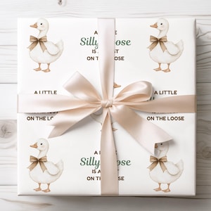 May include: White gift wrap featuring a repeating pattern of a cartoon goose wearing a brown bow tie. The text "A Silly Goose is on the Loose" is printed on the wrap. A light beige ribbon is tied around the gift.