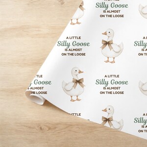 May include: White wrapping paper featuring a repeating pattern of watercolor geese with brown bows. The text "A Little Silly Goose Is Almost On The Loose" is printed in green. The paper is partially rolled.