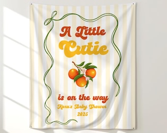 May include: A fabric banner with a vertical striped background in cream and yellow. The banner features the text "A Little Cutie is on the way" in orange and yellow, with a green border and orange illustrations.