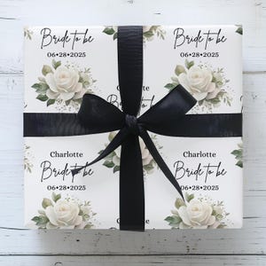 Peut inclure: Papier cadeau blanc avec un motif floral compos&eacute; de roses blanches et de feuilles vertes. Le texte "Bride to be" et la date "06-28-2025" sont r&eacute;p&eacute;t&eacute;s sur le papier d'emballage. Le nom "Charlotte" est &eacute;galement inclus sur le papier d'emballage.