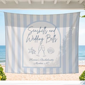 May include: A light blue and white striped beach towel with the text "Seashells and Wedding Bells." The towel features a starfish, champagne glasses, and a seashell graphic. The text "Hanna's Bachelorette Manteo, NC" is also included.