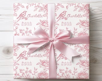 Pode incluir: Papel de embrulho branco com um padrão floral rosa e o texto "Charlotte Bride to be 06-28-2025" repetido quatro vezes. O presente está embrulhado com uma fita de cetim rosa.