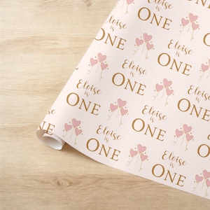 May include: Pink and gold wrapping paper with a repeating pattern of pink heart balloons and the text "Eloise is One".