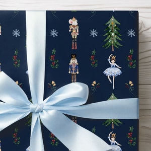 May include: Gift wrapped in navy blue paper with a repeating pattern of nutcrackers, ballerinas, snowflakes, and Christmas trees. A light blue satin ribbon is tied in a bow around the package.