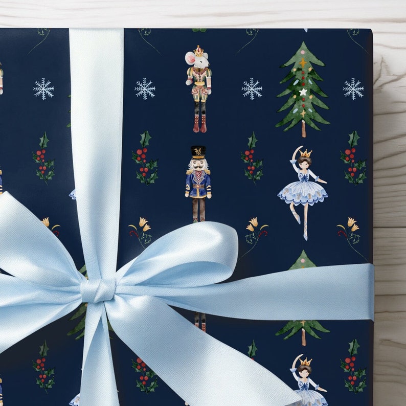 May include: Gift wrapped in navy blue paper with a repeating pattern of nutcrackers, ballerinas, snowflakes, and Christmas trees. A light blue satin ribbon is tied in a bow around the package.