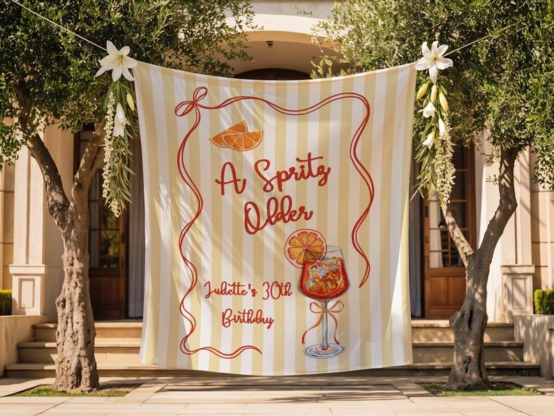 May include: A banner with vertical yellow and white stripes, adorned with the text "A Spritz Older" in red script, along with an illustration of a cocktail and orange slices. The banner also includes the text "Juliette's 30th Birthday".