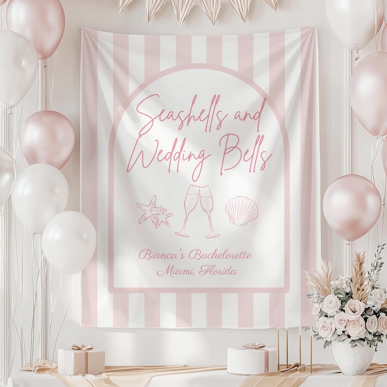 May include: A pink and white striped backdrop with the text "Seashells and Wedding Bells" in a cursive font. The backdrop includes illustrations of a starfish, champagne flutes, and a seashell. Balloons and floral arrangements decorate the scene.