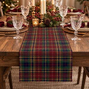 May include: A rectangular table runner featuring a classic plaid pattern in deep red, forest green, navy blue, and thin gold lines. The fabric is laid flat across the center of a wooden dining table, extending from the edge toward the center.