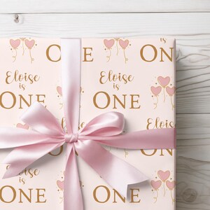 May include: Pink and gold wrapping paper with a pattern of balloons and the text "Eloise is One". A pink ribbon is tied around the paper.