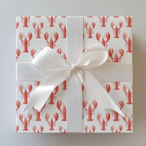 May include: Square gift box wrapped in white paper with a repeating pattern of red lobsters. A wide white satin ribbon is tied around the box with a large bow on top. The box is presented on a light gray background.