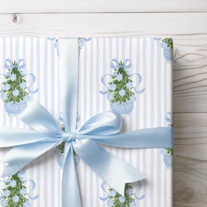 May include: A wrapped gift with light blue satin ribbon. The wrapping paper features a repeating pattern of Christmas trees in blue pots on a white background with vertical blue stripes. The ribbon is tied in a bow.