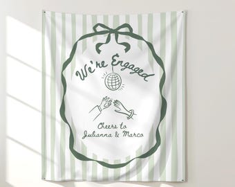 May include: A white and green striped tapestry with a green ribbon border. The text "We're Engaged" is written in a script font, along with the names "Julianna & Marco". A drawing of hands exchanging a ring is also included.