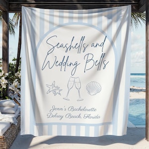May include: A beach towel with a white and pale blue striped design. The towel has the text "Seashells and Wedding Bells" and illustrations of a starfish, champagne flutes, and a seashell. The text "Jenn's Bachelorette Delray Beach, Florida" is also included.