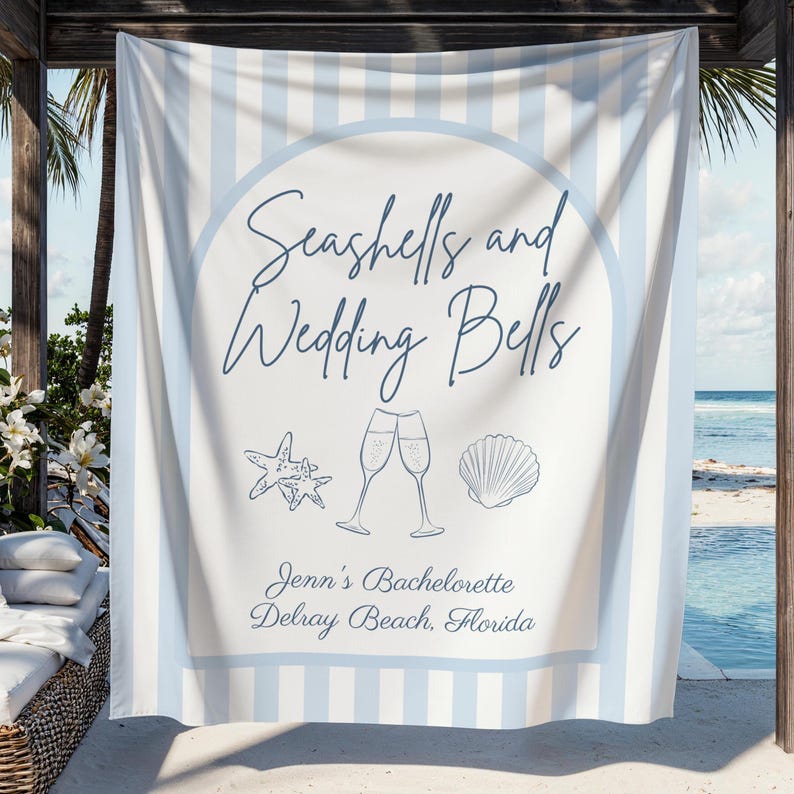 May include: A white and light blue striped beach towel with the text "Seashells and Wedding Bells." The towel features illustrations of a starfish, champagne flutes, and a seashell. The text "Jenn's Bachelorette Delray Beach, Florida" is also included.