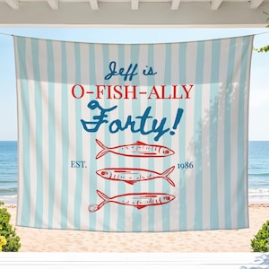 May include: A rectangular fabric banner featuring a light blue and white vertical striped pattern. The text reads "Jeff is O-FISH-ALLY Forty!" in blue and red, with "EST. 1986" and three red fish illustrations centered below the text.