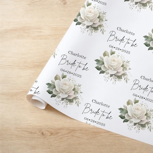 Peut inclure: Papier d'emballage floral blanc avec un motif r&eacute;p&eacute;t&eacute; de roses blanches et de feuilles vertes. Le texte "Charlotte Bride to be 06-28-2025" est r&eacute;p&eacute;t&eacute; sur le papier.