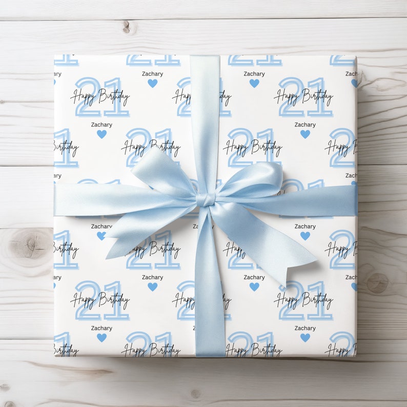 May include: White gift wrap with a blue and white pattern. The pattern features the number 21 with the words "Happy Birthday" and the name "Zachary" repeated throughout. A blue ribbon is tied around the gift box.