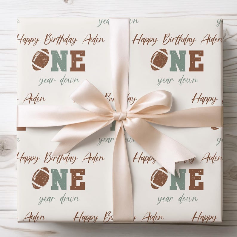 Op de afbeelding: Witte cadeaupapier met een bruin en groen voetbalontwerp. De tekst "Happy Birthday Aiden One year down" wordt herhaald op het cadeaupapier.