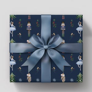 May include: A wrapped gift with navy blue wrapping paper featuring a pattern of nutcrackers, ballerinas, and Christmas trees. A wide, light blue satin ribbon is tied in a large bow around the gift.