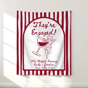 May include: A rectangular wall tapestry featuring vertical dark red and white stripes. The center displays an arched frame containing the text They're Engaged!, a line drawing of a cherub holding a wine glass, and the text The Perfect Pairing Emily + Jordan Napa Valley 2026.