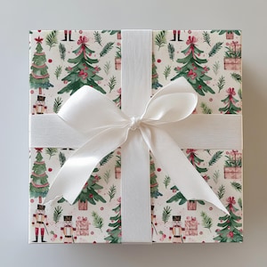 Puede incluir: Una caja de regalo cuadrada envuelta en papel con temática navideña. El papel presenta un patrón de árboles de Navidad verdes, cascanueces y regalos sobre un fondo crema. Una cinta ancha de satén blanco está atada con un lazo en la parte superior.