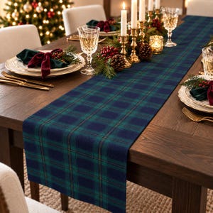 May include: A long rectangular table runner featuring a dark blue and green plaid pattern. The runner is centered on a wooden table set with white plates, gold-toned cutlery, crystal glassware, and brass candle holders with white candles.
