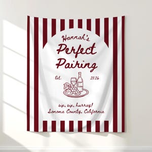 May include: A white wall tapestry with a burgundy striped border and a central design. The design features the text "Hannah's Perfect Pairing" and an illustration of wine, grapes, and cheese. The text "Est. 2026" is also present.