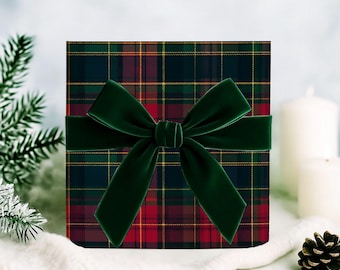 May include: A square gift box with a tartan pattern in red, green, and yellow. A dark green velvet ribbon is tied around the box in a bow. The box is on a white surface with a pine cone and candles in the background.