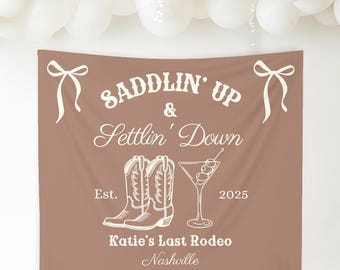 Peut inclure: Une tapisserie brune avec du texte blanc qui dit "Saddlin' Up & Settin' Down" avec une paire de bottes de cowboy, un verre à martini et le texte "Katie's Last Rodeo Nashville Est. 2025".