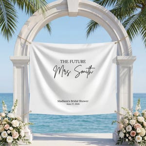 May include: A white banner with the text "THE FUTURE Mrs. Smith" in black script, displayed on a white archway. The banner also includes "Madison's Bridal Shower June 27, 2026". The scene is set by the ocean with floral arrangements.