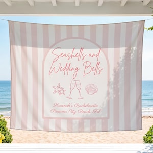 May include: A pink and white striped beach towel with the text "Seashells and Wedding Bells" in a decorative font. The towel features a seashell and champagne glass graphic, with the text "Hannah's Bachelorette Panama City Beach, FL".