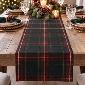 Puede incluir: Un camino de mesa rectangular con un patrón de cuadros en verde oscuro y rojo intenso con líneas doradas finas. La tela tiene una textura tejida y se extiende por el centro de una mesa de comedor de madera, entre los cubiertos.